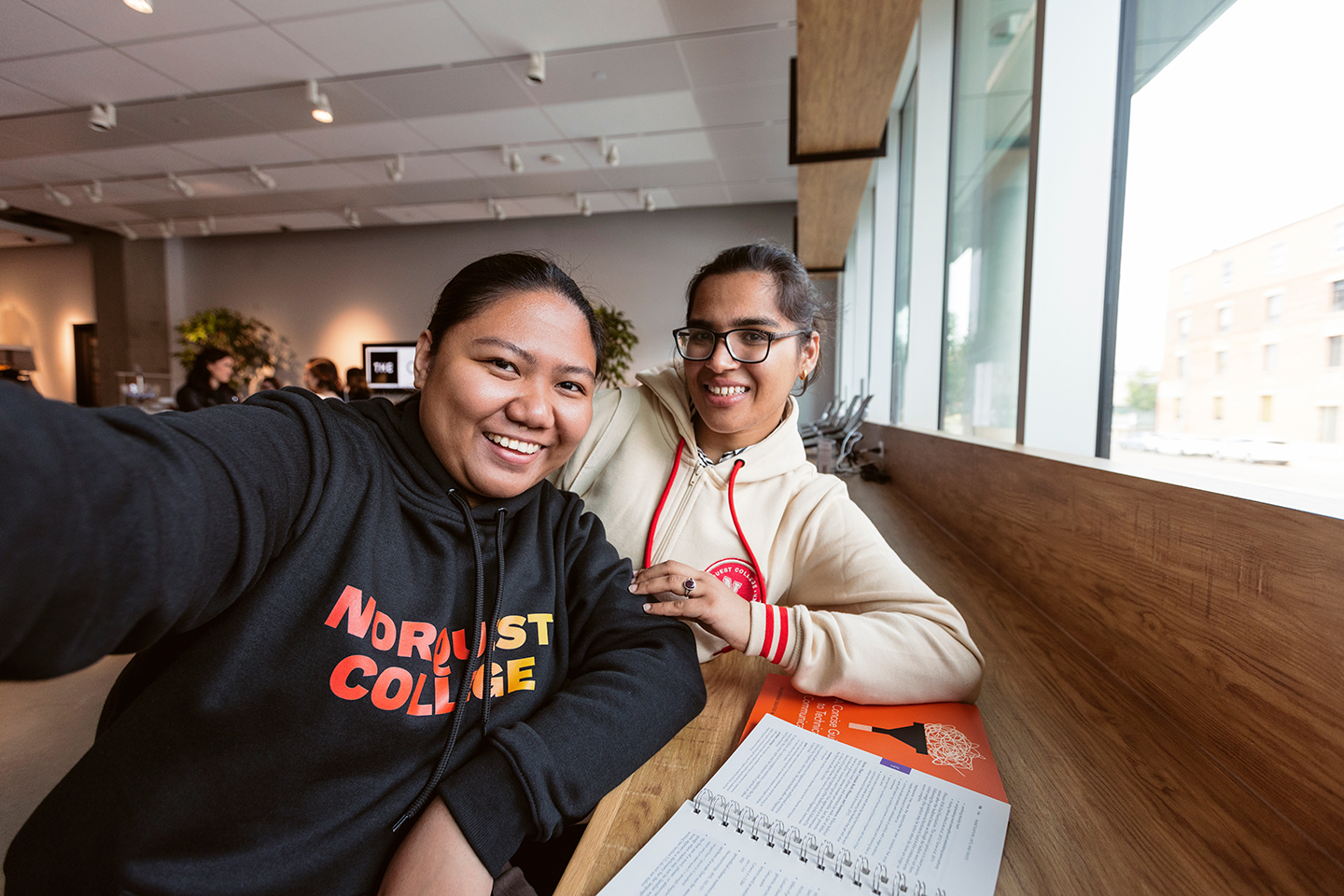Two female students leaning against a long desk taking a selfie in the core