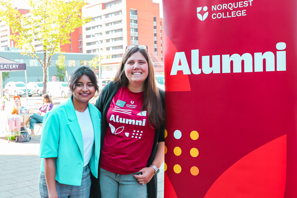 Two smiling women standing in front of a NorQuest College Alumni banner